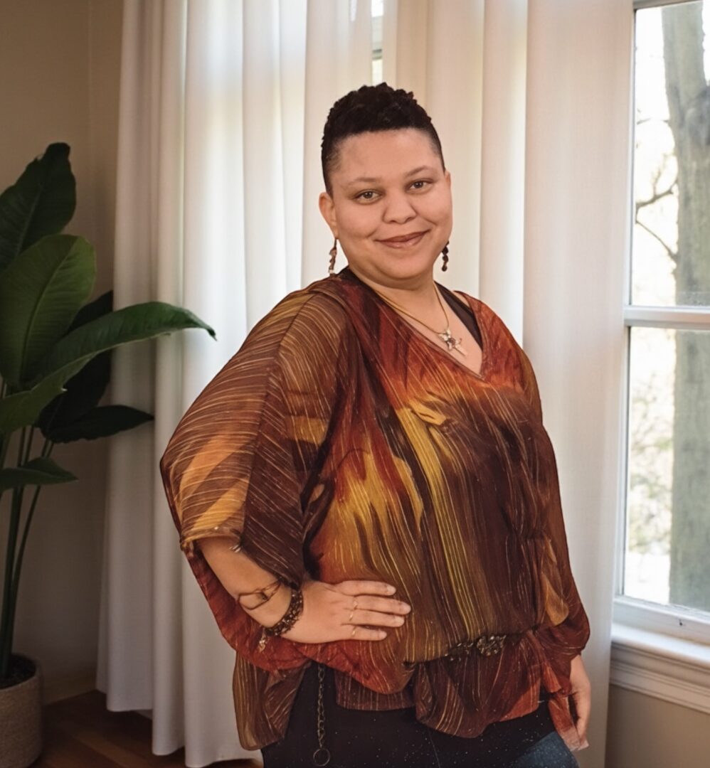 Woman with short hair wearing a colorful, loose blouse, standing indoors near a window with curtains.