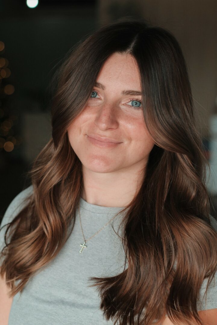 Young woman with long, wavy brown hair and blue eyes smiling, wearing a light gray shirt and necklace.