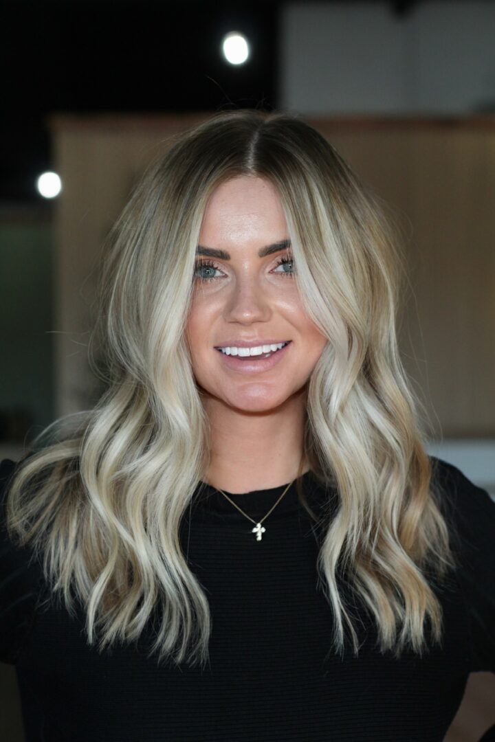 Young woman with long wavy blonde hair smiling, wearing a black top and a cross necklace.