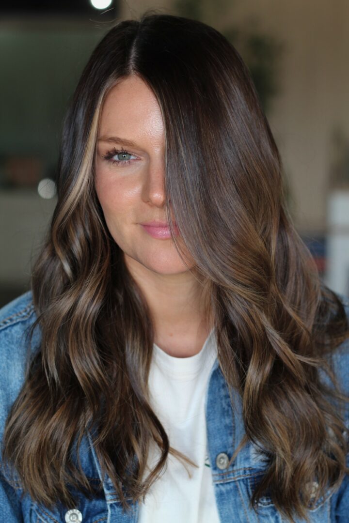 Woman with long wavy brown hair, wearing a white shirt and denim jacket, looking to the side.