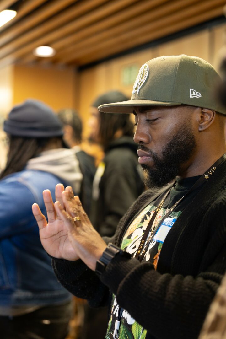 Man with a beard wearing a green cap and black jacket, focusing on his hands in a room with people.