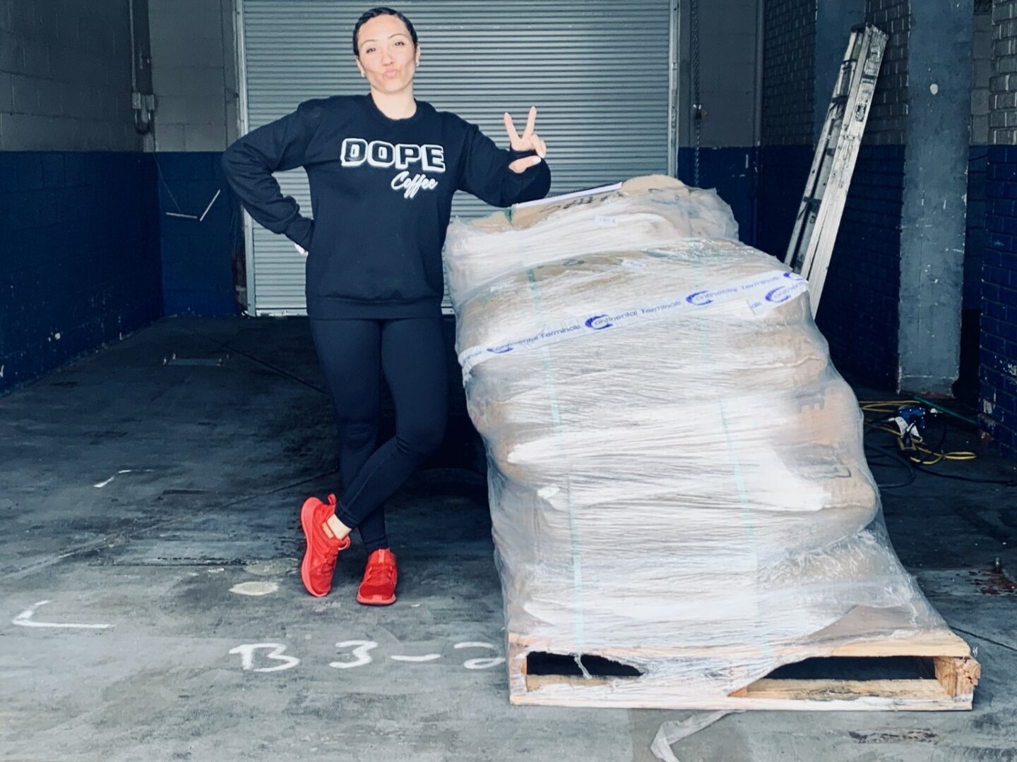 Woman standing next to a large pallet wrapped in plastic in an industrial space, making a peace sign with her right hand.
