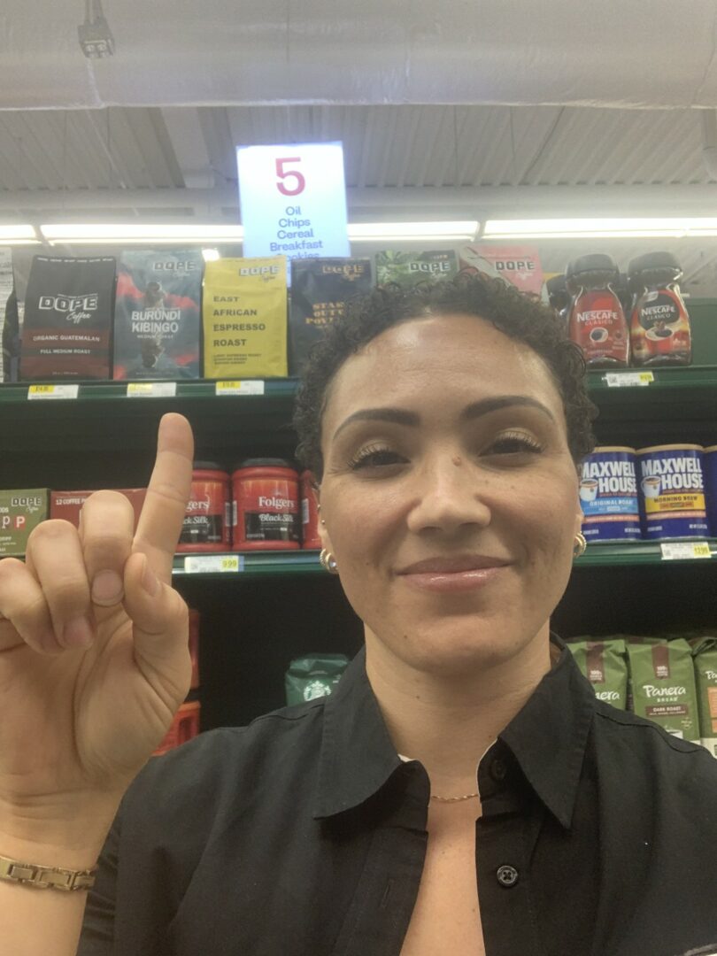 Woman with dark hair smiling, holding up her index finger in a store aisle with shelves of products behind her.