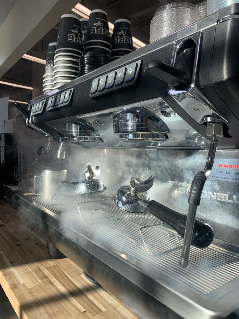 Espresso machine with steam rising, cups on top, and portafilters attached, in a coffee shop setting.