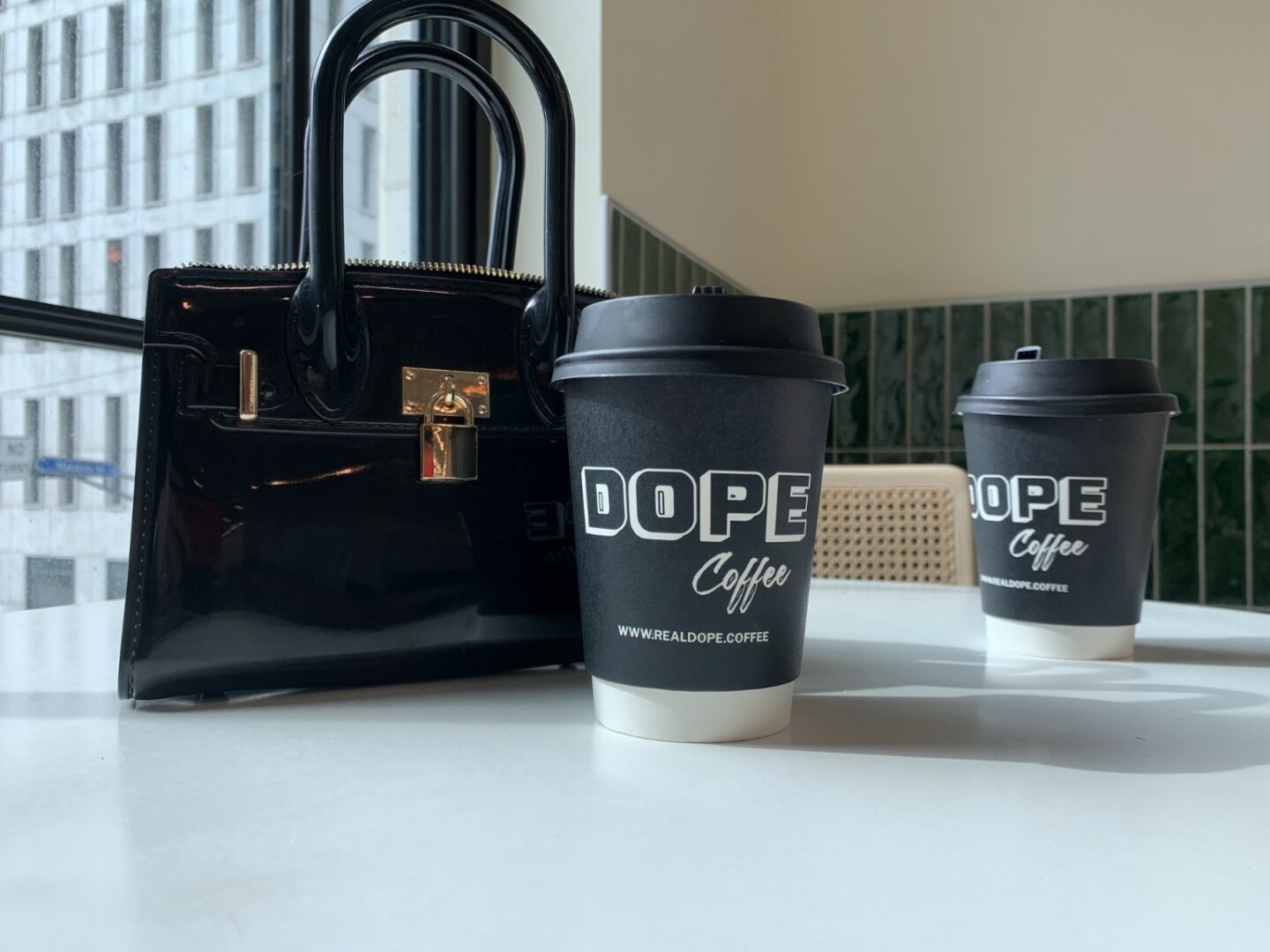 Black handbag and two coffee cups on a white table near a window with city buildings outside.