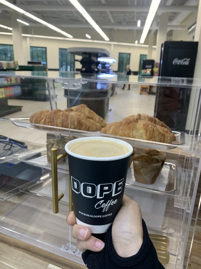 Hand holding a coffee cup in front of a display case with croissants inside a cafe or store.