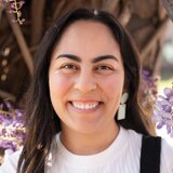Smiling woman with long dark hair, wearing white earrings and a white top, outdoors with purple flowers in background.
