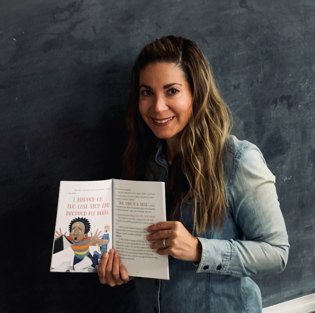 Woman with long wavy hair holding an open book, smiling, standing against a dark wall.