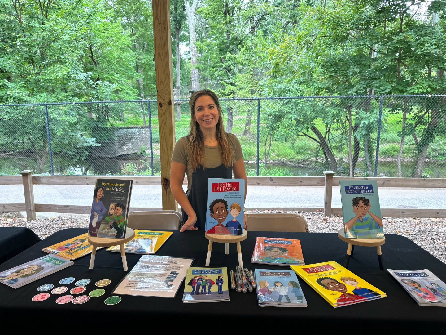 Woman standing behind a table with books and promotional materials outdoors, trees and a fence in background.
