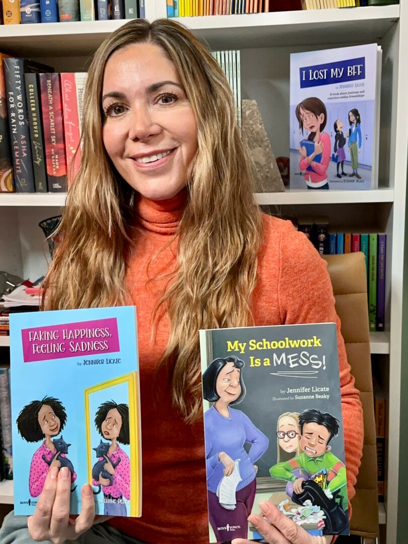 Woman with long wavy hair smiling, holding two children's books, in front of a bookshelf.
