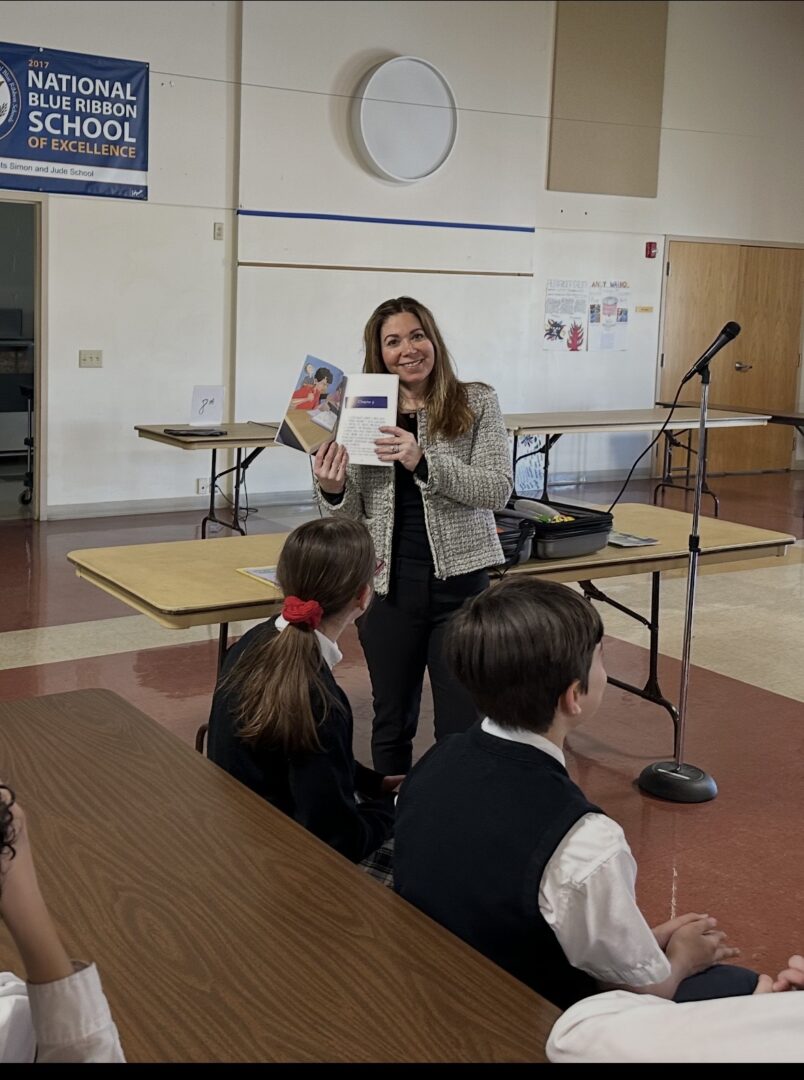 Woman standing at front of classroom holding a book, students seated at desks listening, classroom with presentation equipment.
