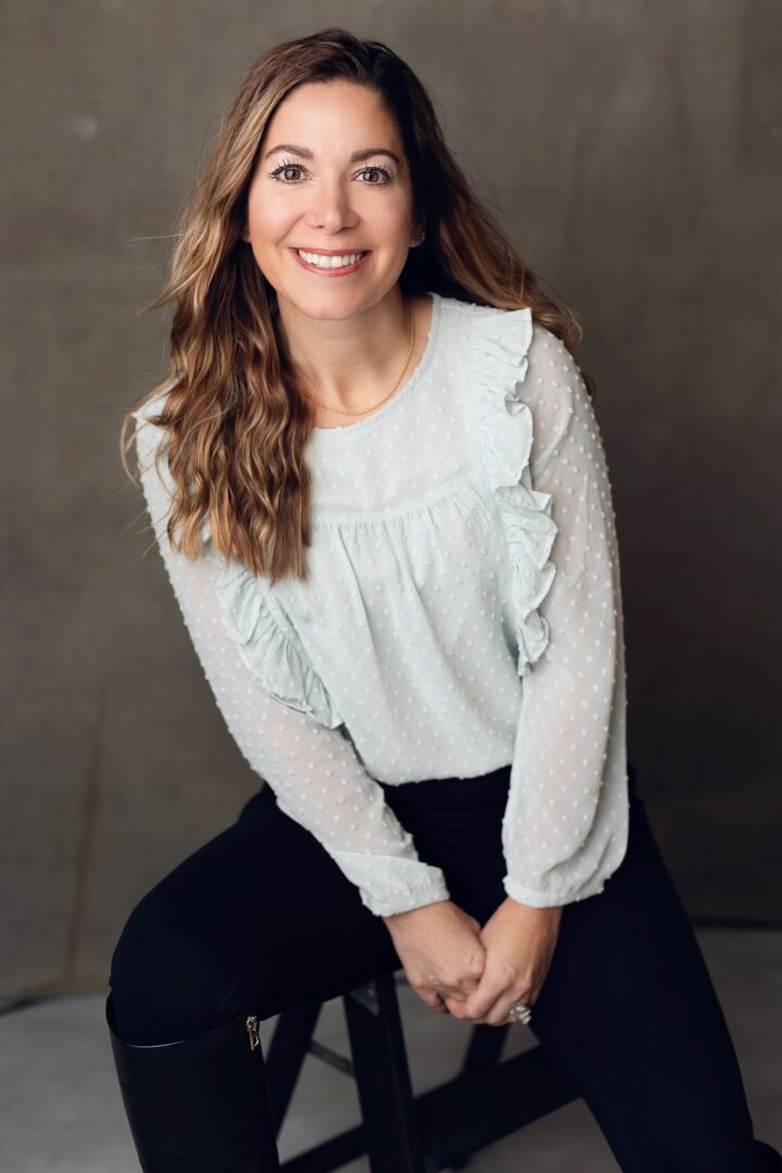 Woman with long wavy hair smiling, sitting on a stool, wearing a white blouse and black pants.