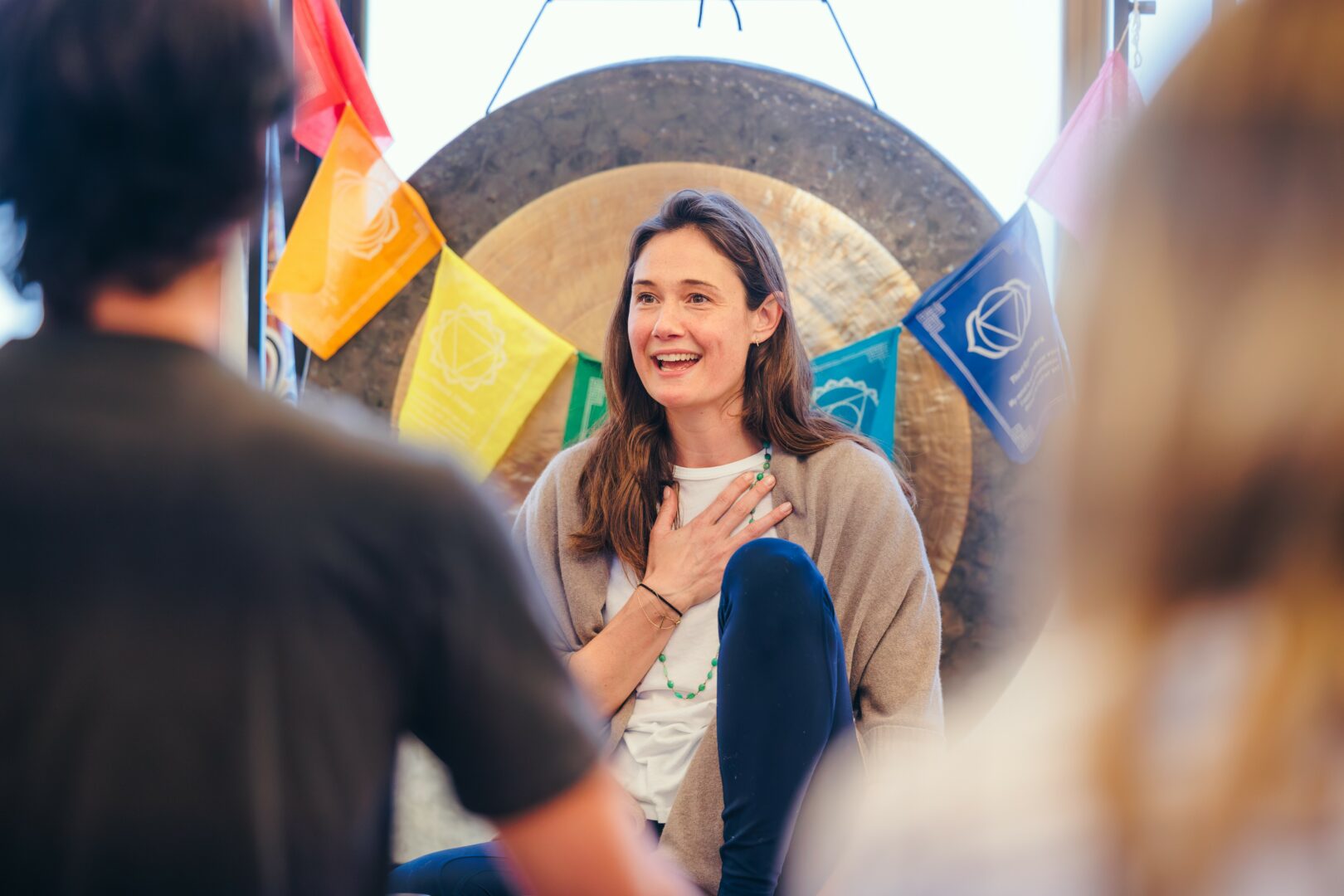 Woman smiling with hand on chest, surrounded by people, colorful flags, and a large gong in background.