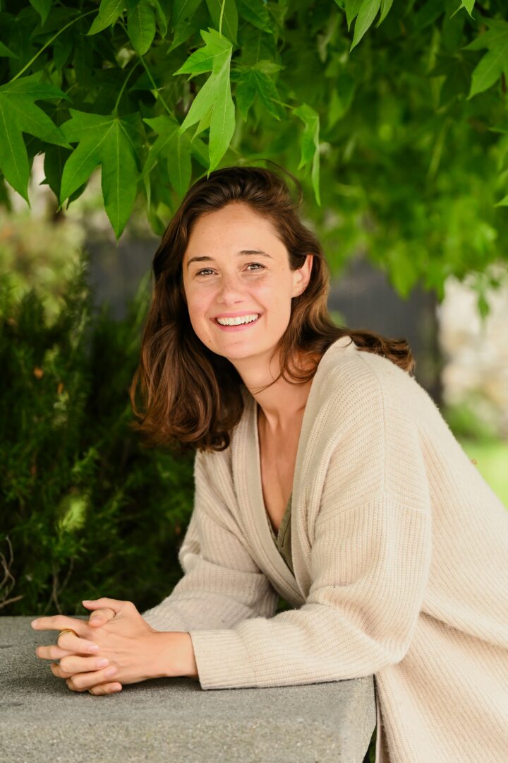 Smiling woman with brown hair in a beige sweater outdoors, surrounded by green leaves and trees.