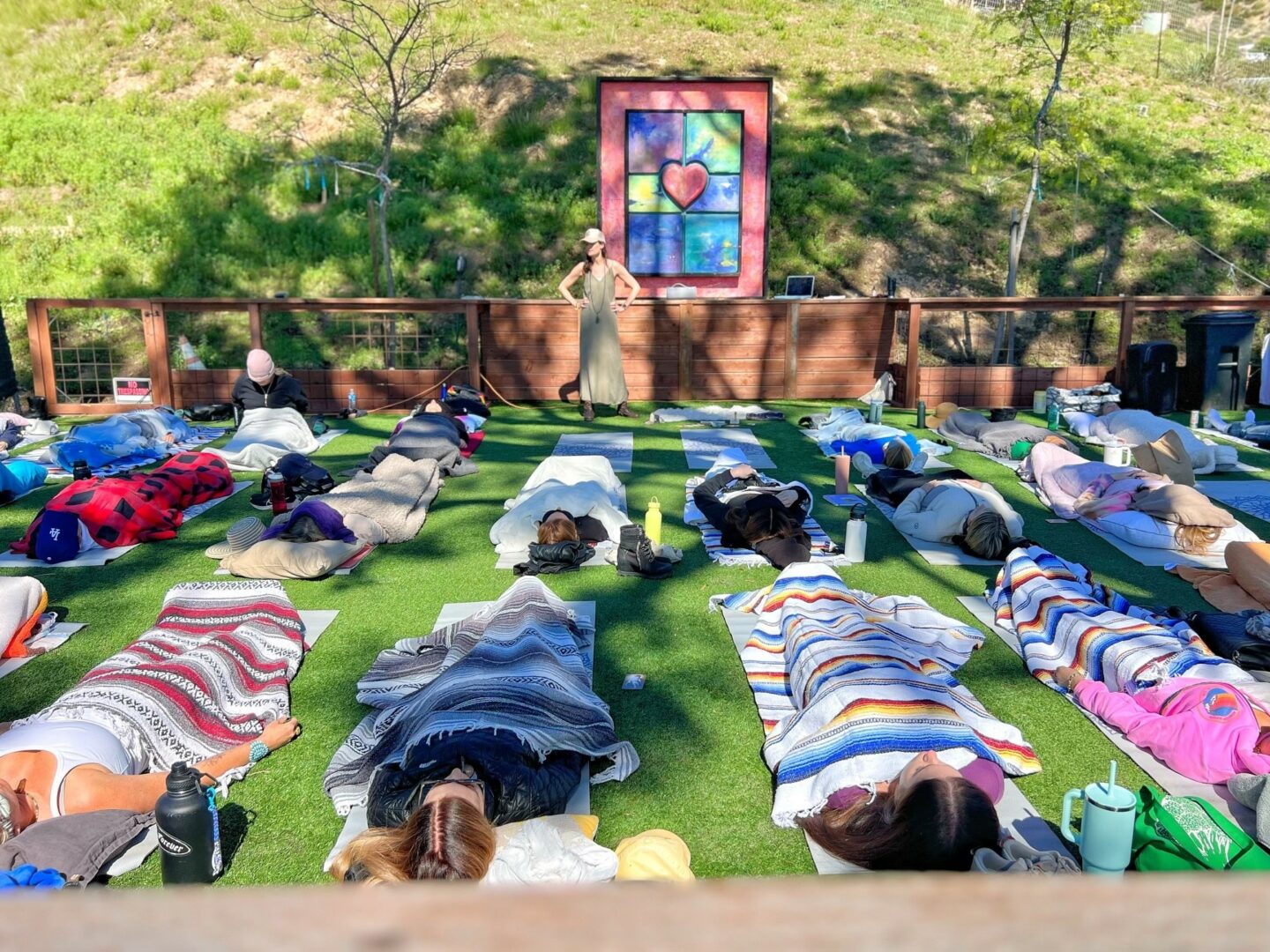 People lying on blankets outdoors with a woman standing near a wooden fence and a colorful stained glass window in the background.