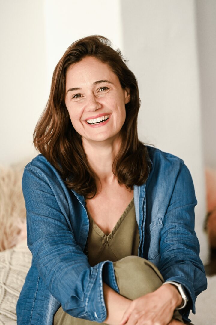 Smiling woman with shoulder-length brown hair wearing a green top and denim jacket indoors.