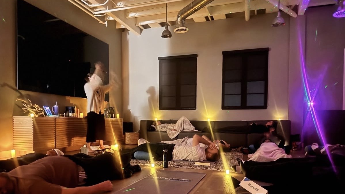 People in a dimly lit room with couches, a large TV, and colorful lights, some lying down and one standing near the TV.