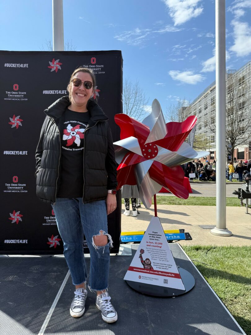 Person standing outdoors near a display with a large red and white windmill decoration, black backdrop, and informational sign.