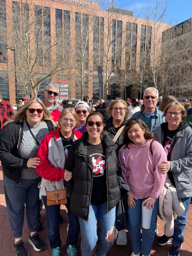 Group of people standing outdoors in front of a brick building with trees, some wearing sunglasses and casual clothing.