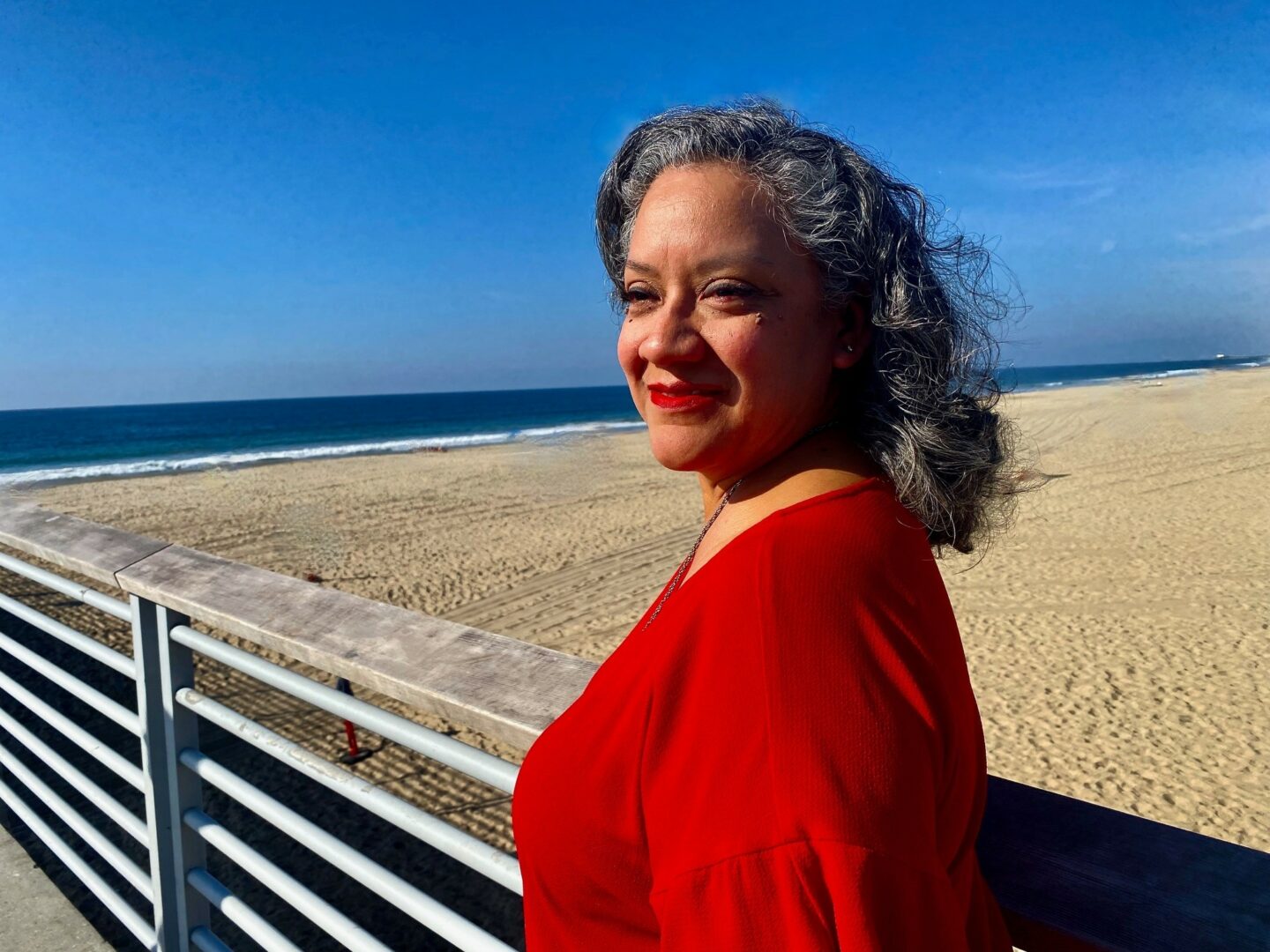 Woman with curly gray hair and red lipstick smiling at the beach, wearing a red top, with sand and ocean in background.