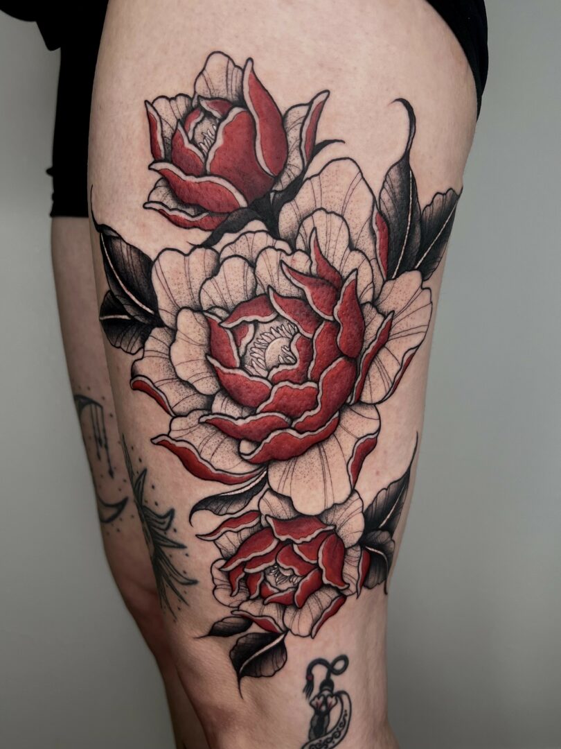Tattoo of large red and beige flowers with black leaves on person's thigh.