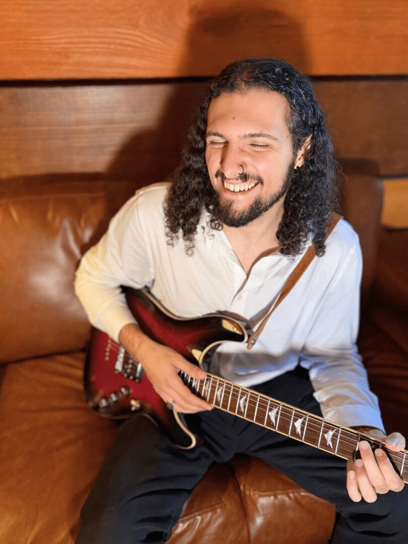 Person with long curly hair smiling while playing an electric guitar on a leather couch.