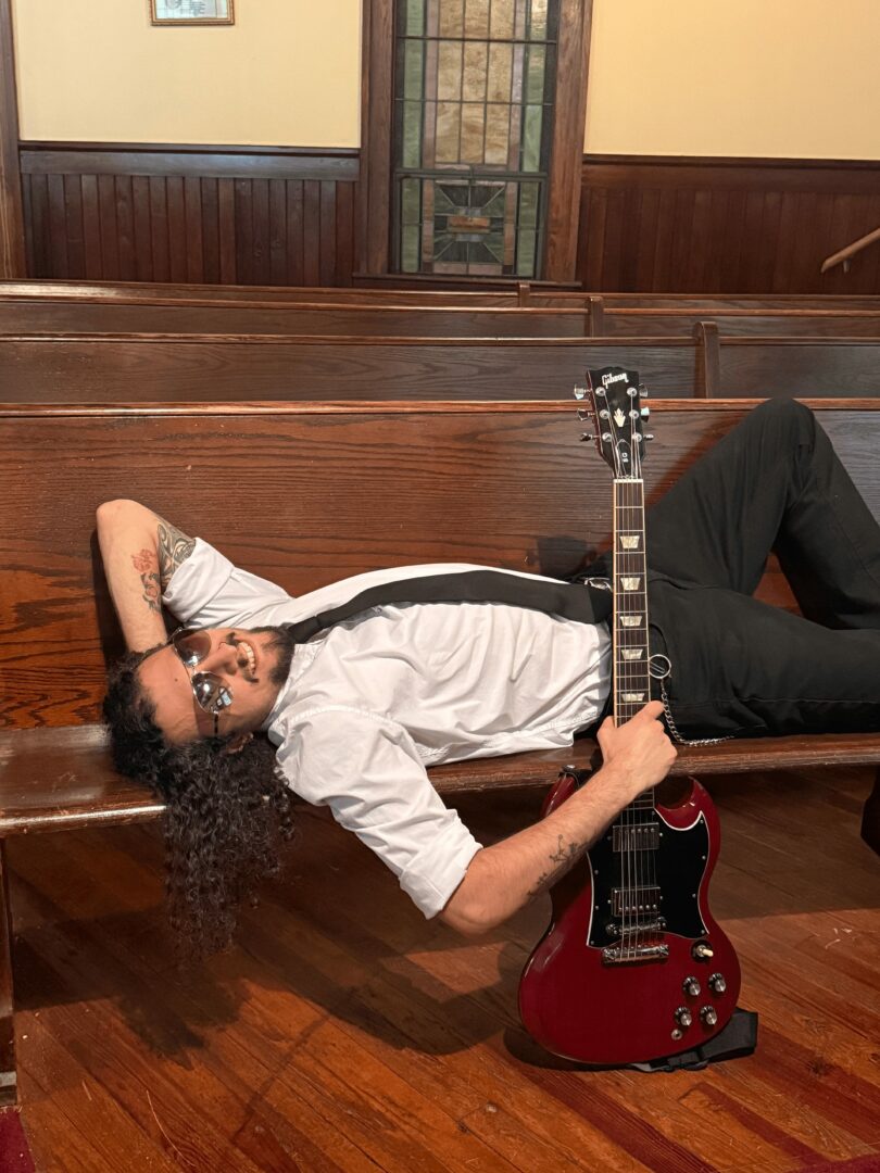 Person lying on wooden floor with guitar, in a room with wooden pews and stained glass window behind.