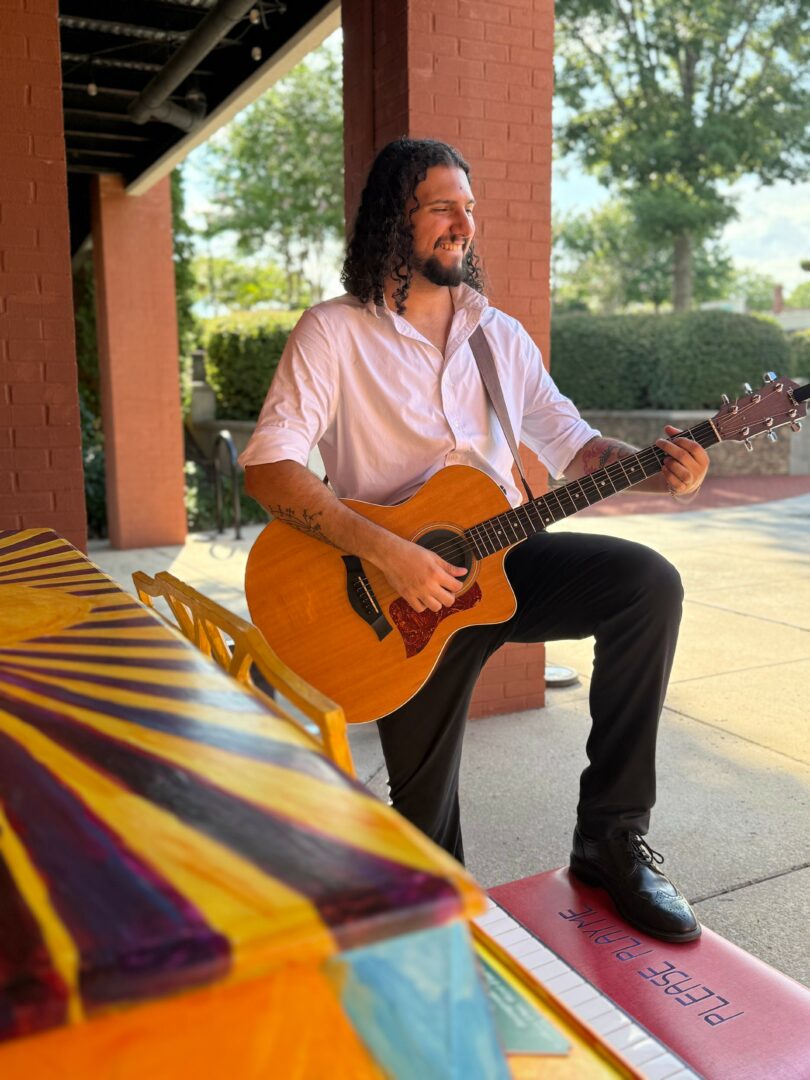 Man with long hair and beard playing an acoustic guitar outdoors near a brick structure and trees.