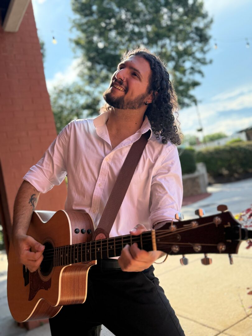 Man with long curly hair playing an acoustic guitar outdoors, smiling, with trees and a building in the background.