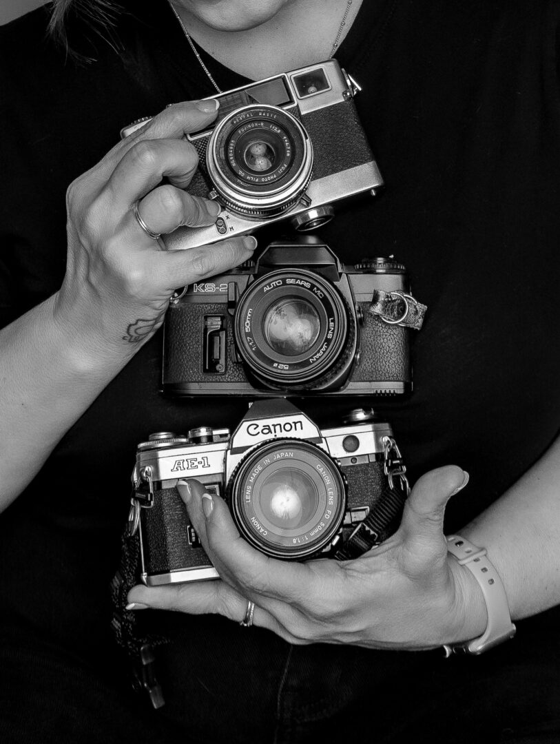 Person holding three cameras, one in each hand and one hanging from neck, black and white photo.