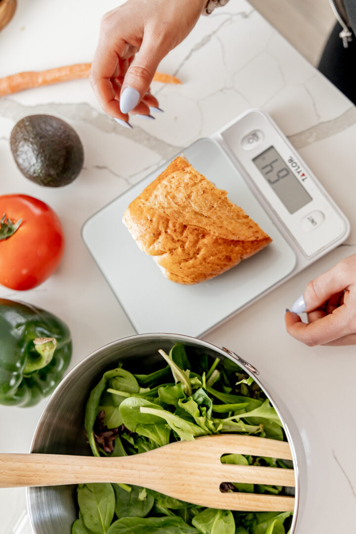 Person's hand weighing a piece of bread on a digital kitchen scale with vegetables nearby.