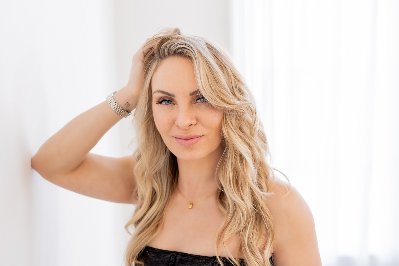 Woman with long wavy blonde hair, smiling, wearing a black strapless top, standing indoors near a white curtain.