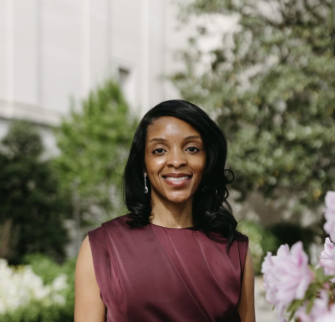Smiling woman with black hair in a sleeveless maroon dress outdoors with greenery and flowers.