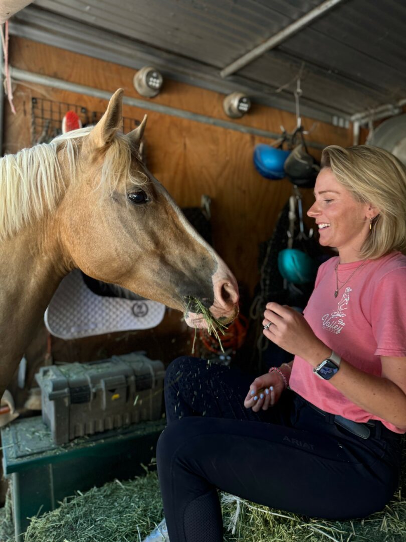 Woman in pink shirt smiling at a light-colored horse inside a stable.