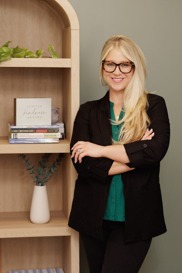 Woman with long blonde hair, glasses, black blazer, and green top standing with arms crossed beside a bookshelf.