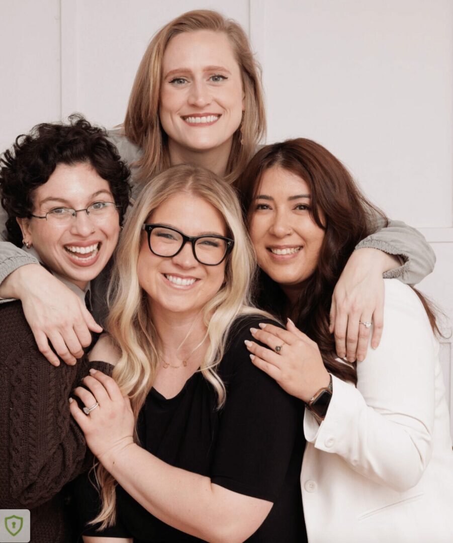 Four women smiling, hugging, and posing together indoors.