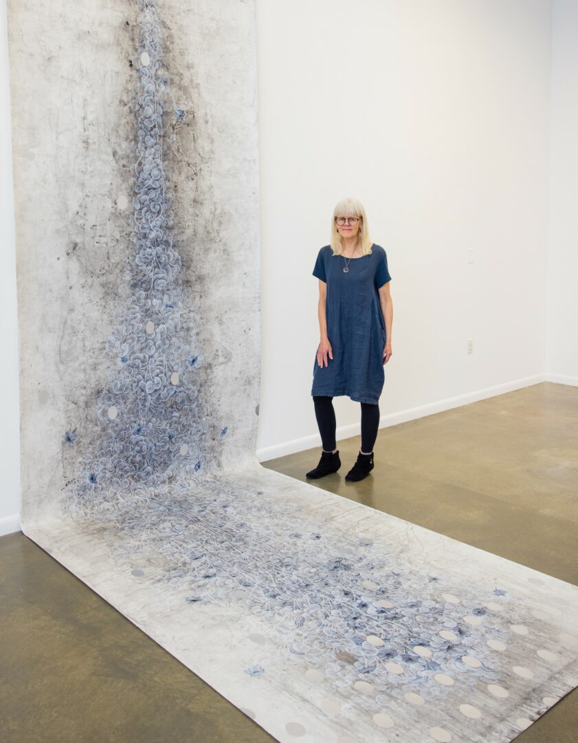 Woman with glasses and blonde hair stands in art gallery next to large textured artwork on the floor and wall.