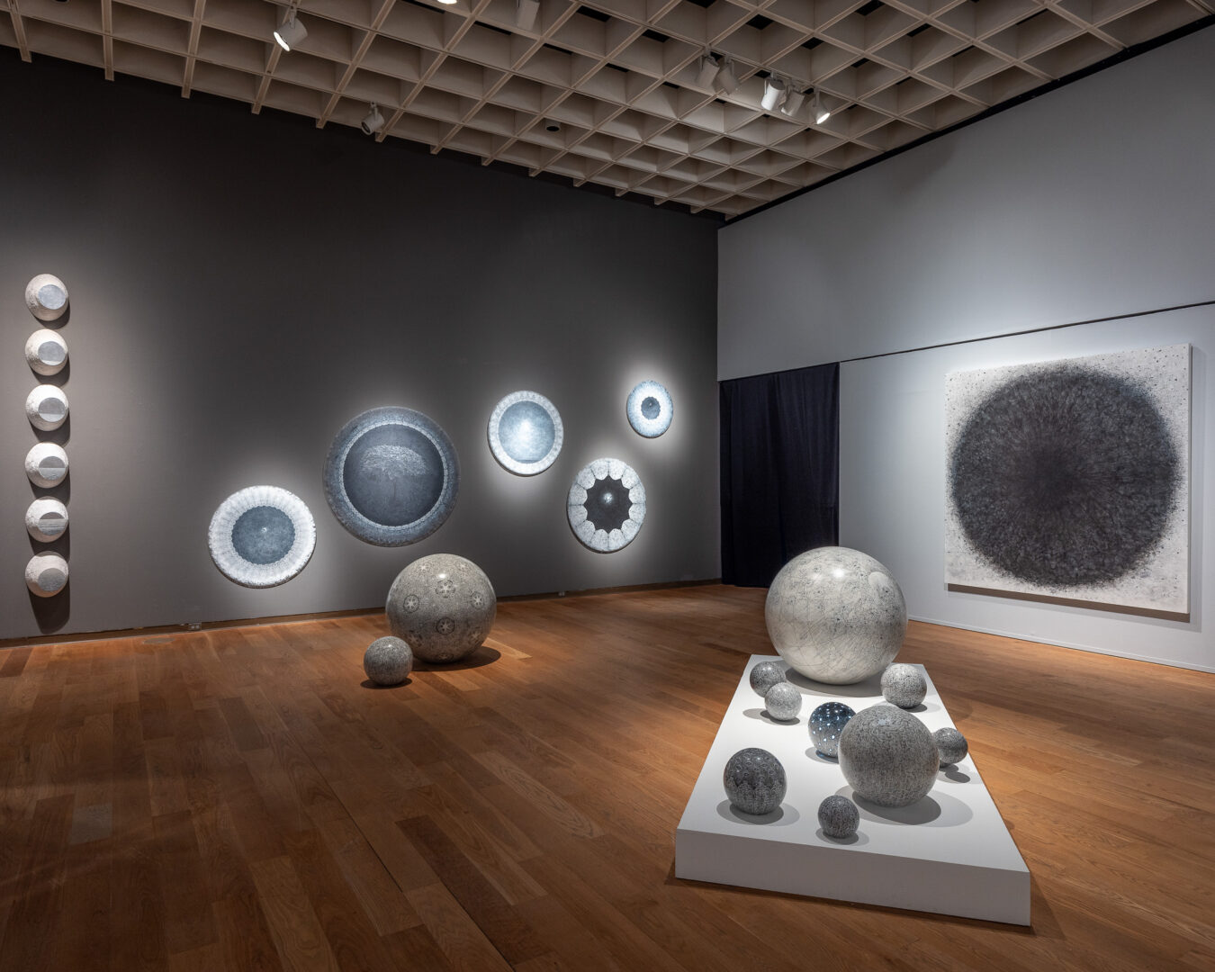Art gallery with circular and spherical sculptures and wall-mounted circular art pieces, wooden floor, and ceiling grid.