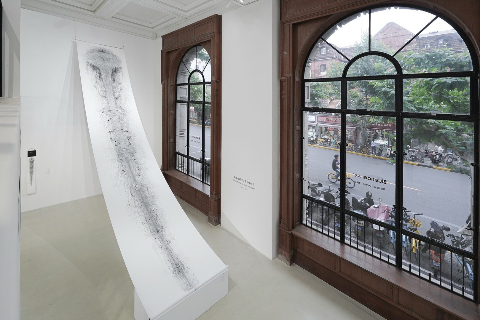 White sculpture with black markings on a pedestal in a gallery with large arched windows showing a street scene.