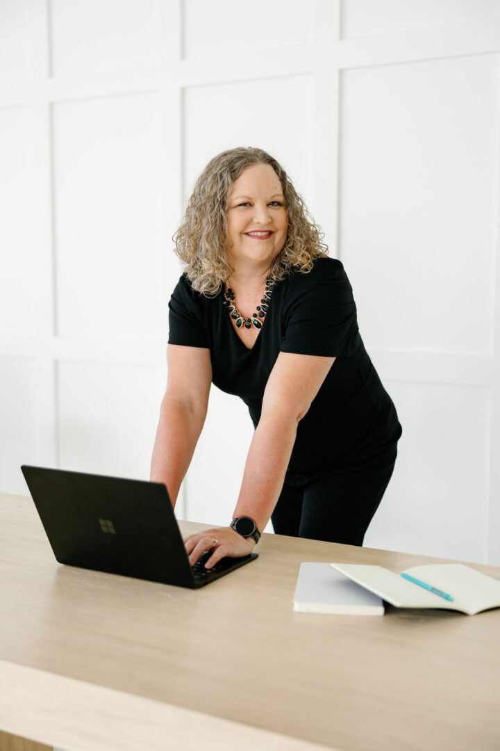 Woman with curly hair in black dress leaning on a table with a laptop and open book.