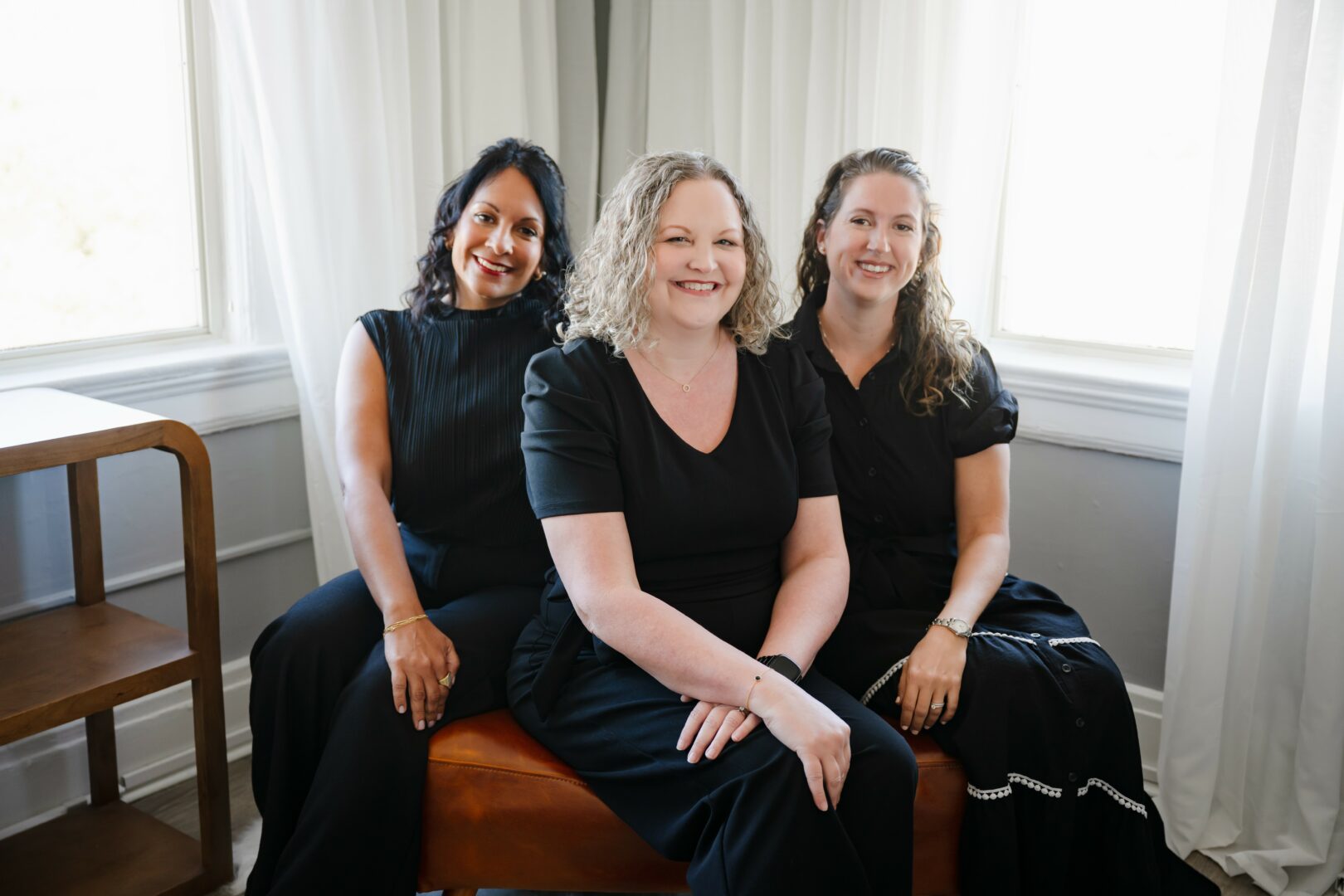 Three women sitting on a bench in a bright room with large windows and white curtains.