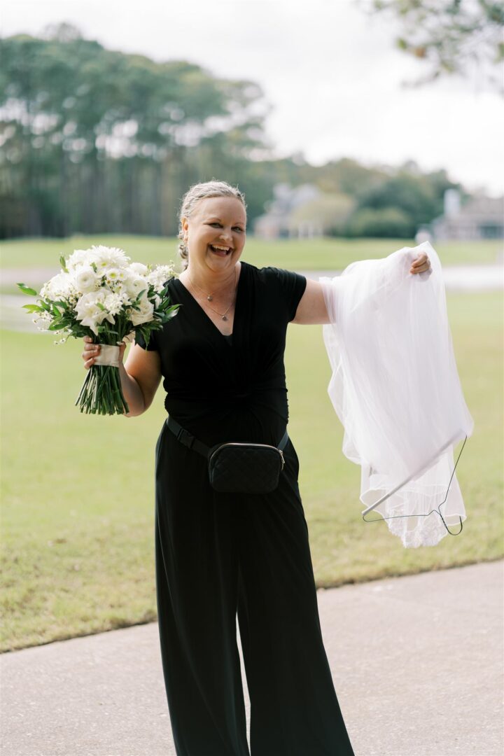 Woman in black dress holding bouquet and a white cloth outdoors, smiling, with trees and grass in background.