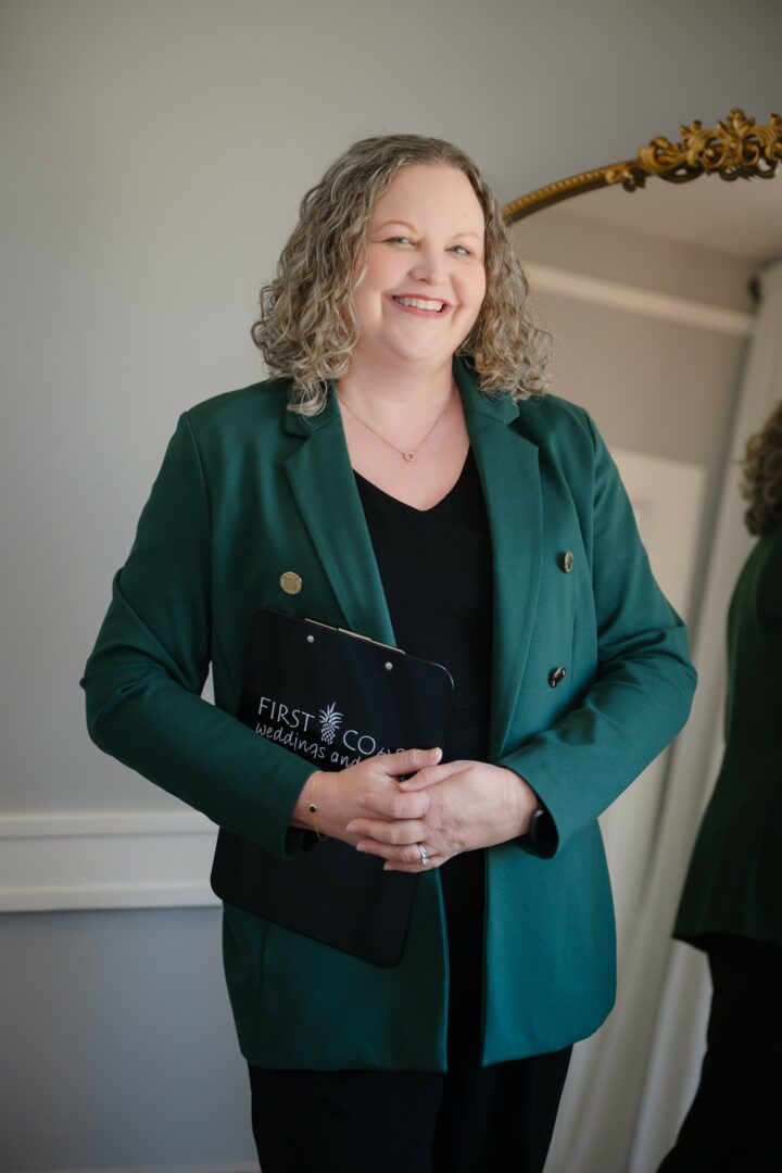 Woman with curly hair smiling, holding a black folder, wearing a green blazer and black top, standing indoors.